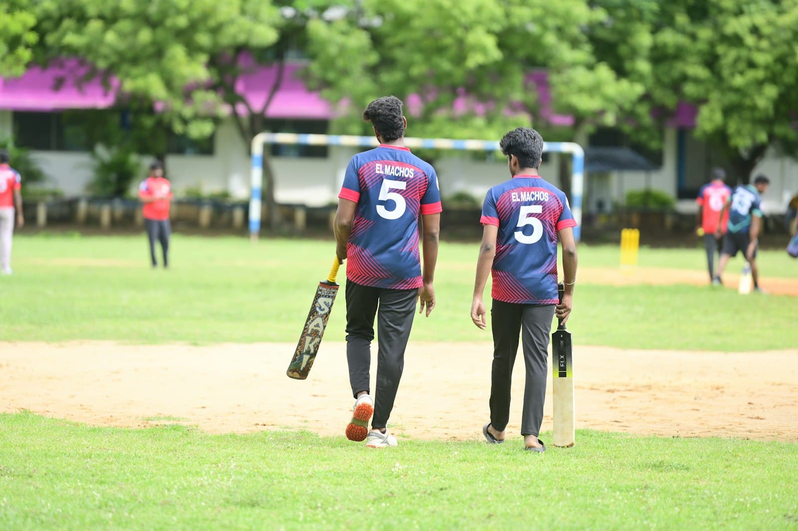 Two teammates in number five shirts walking across a cricket ground with their bats