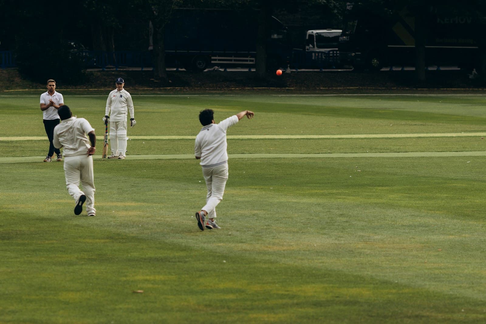 A bowler lets go of a red ball on a green club ground while fielders watch