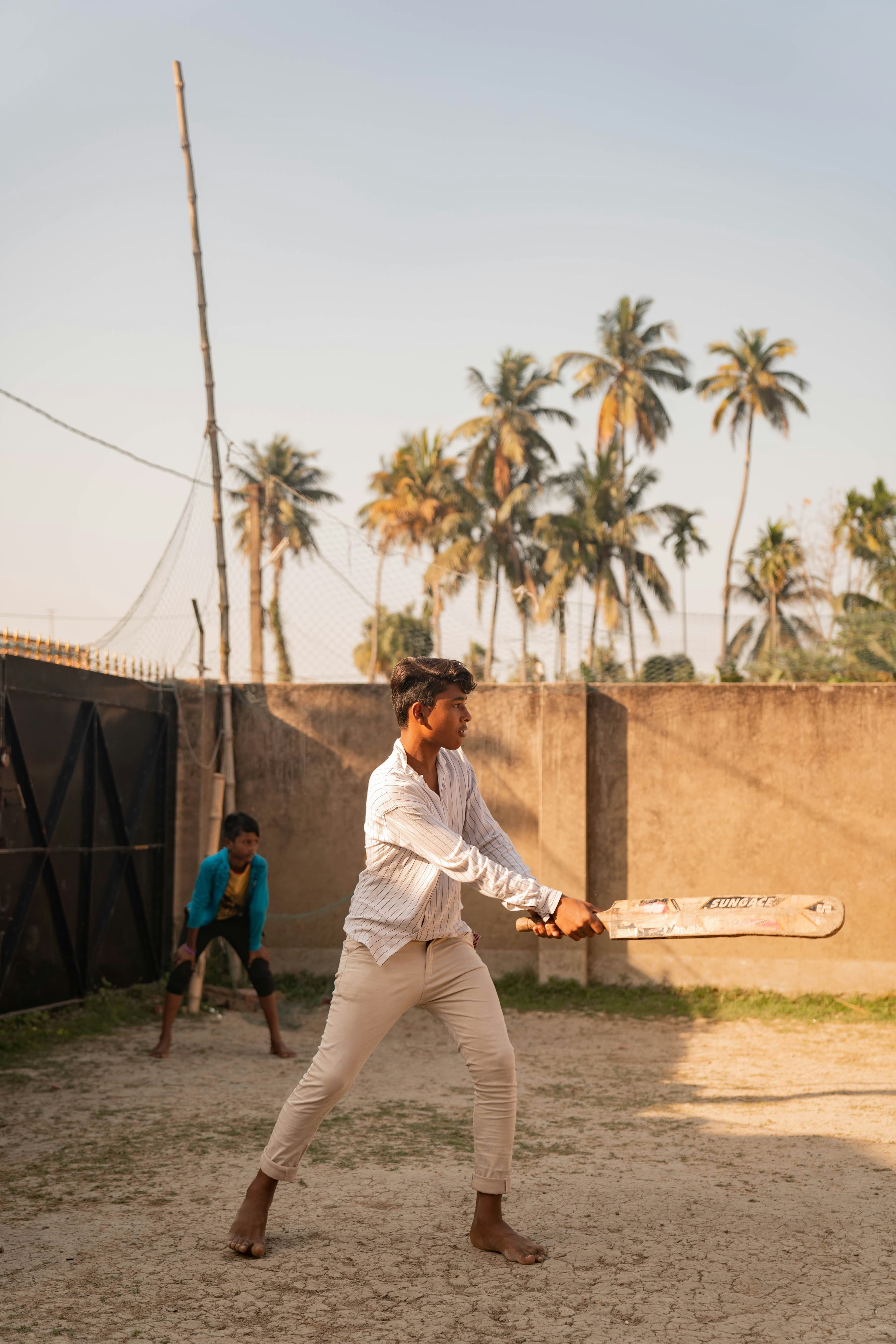 A batter mid-swing on a sandy ground lined with palm trees