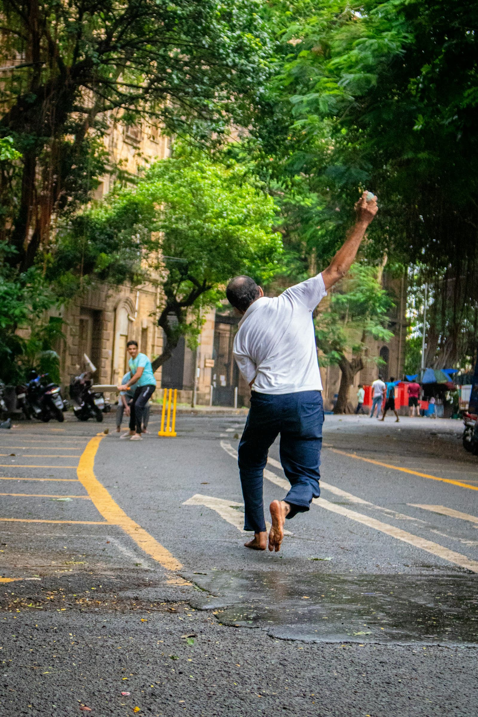 A bowler mid-delivery on a tree-lined street, the batter waiting at the far end